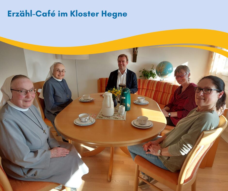 Um einen runden hölzernen Tisch sitzen fünf Menschen und trinken Kaffee