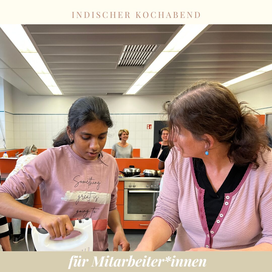 zwei Frauen in einer großen Küche, die eine hält ein Rührgerät in der Hand. Im Hintergrund sind andere Menschen am arbeiten.