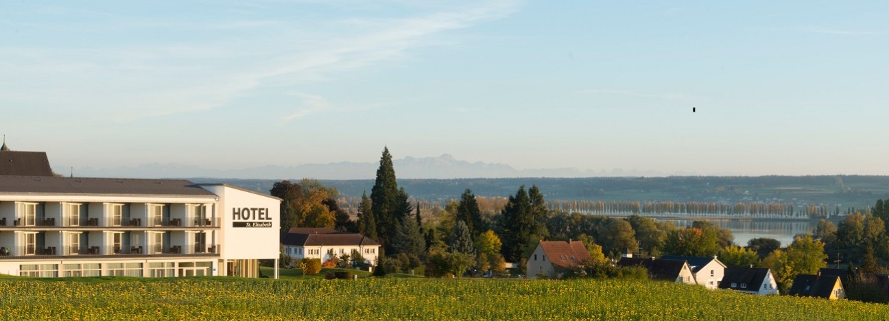 A section of the Hotel St.&nbsp;Elisabeth, with a field in the foreground and Lake Constance and mountains in the background