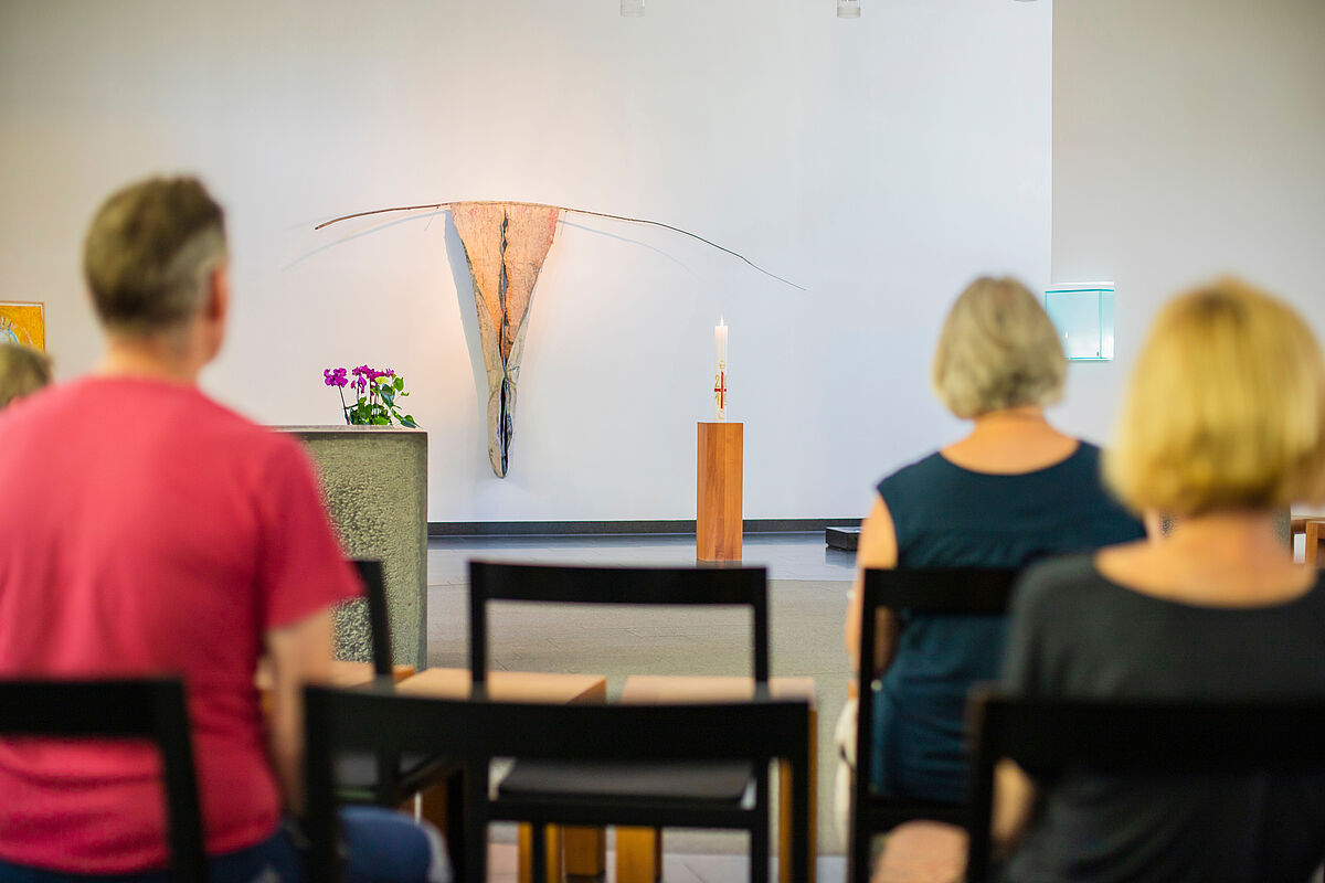 Drei Menschen von hinten, die in einer bestuhlten Kirche sitzen. Neben dem Altar brennt eine weiße Kerze.